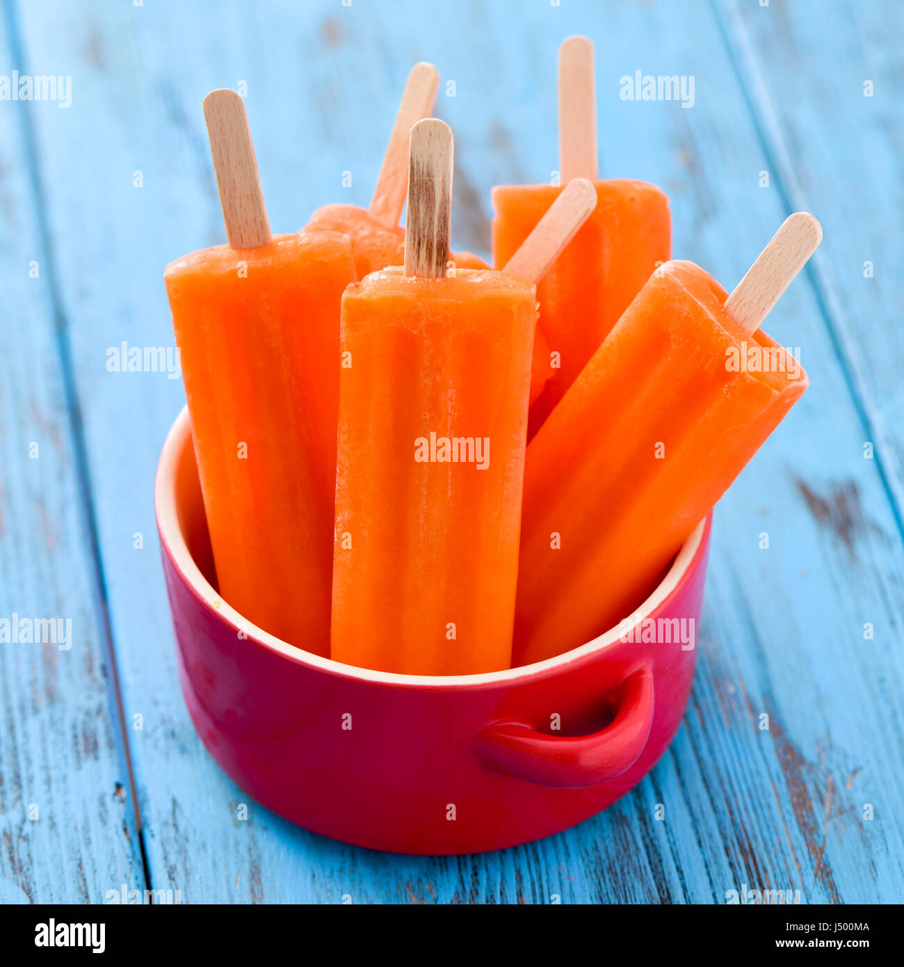 einige erfrischende orange aromatisierte Eis am Stiel in einem roten Keramikschale auf einem blauen rustikalen Holztisch Stockfoto