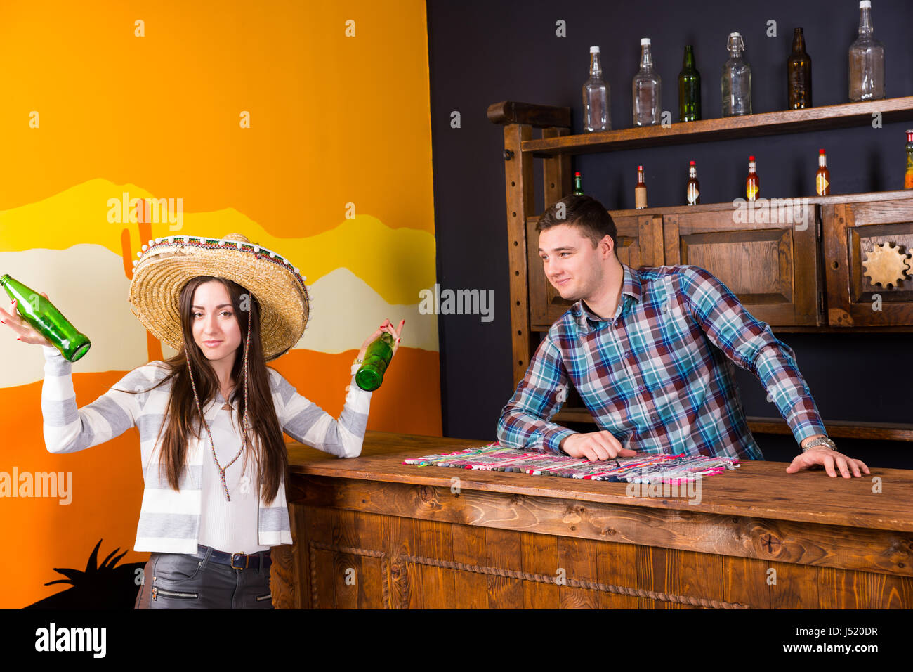 Eine junge Frau in einen Sombrero halten zwei Flaschen Bier und stehen an der Bar neben der Barkeeper in einem karierten Hemd in einer mexikanischen bar Stockfoto