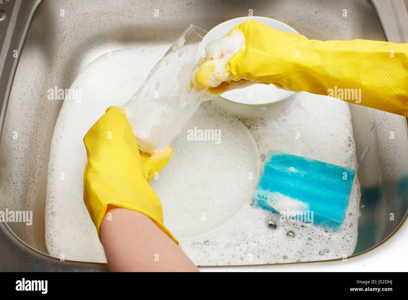Nahaufnahme von weiblichen Händen schützende Gummihandschuhe Trinkglas mit blauen Reinigung Waschen gegen Küchenspüle voller Schaum und Registerkarte "sponge gelb Stockfoto