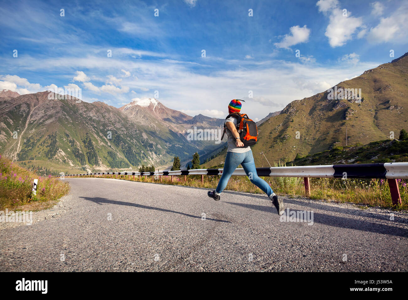 Tourist-Frau mit Rucksack und Regenbogen Hut liefen die Asphaltstraße in den Bergen in der Nähe von Big Almaty-See in Kasachstan Stockfoto