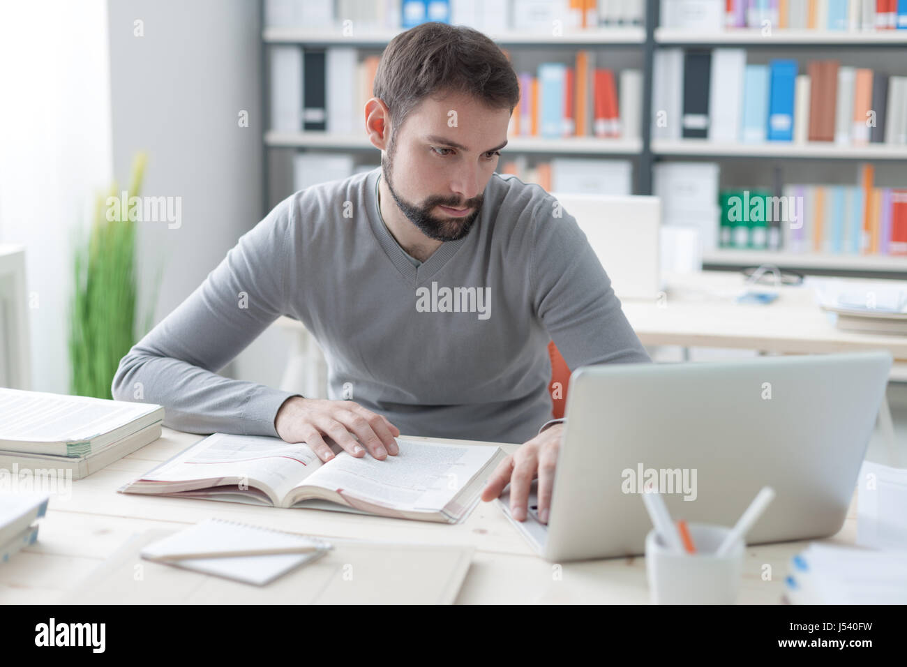 Junger hübscher Mann am Schreibtisch sitzen, ein Buch und Verbindung zum Internet mit einem laptop Stockfoto