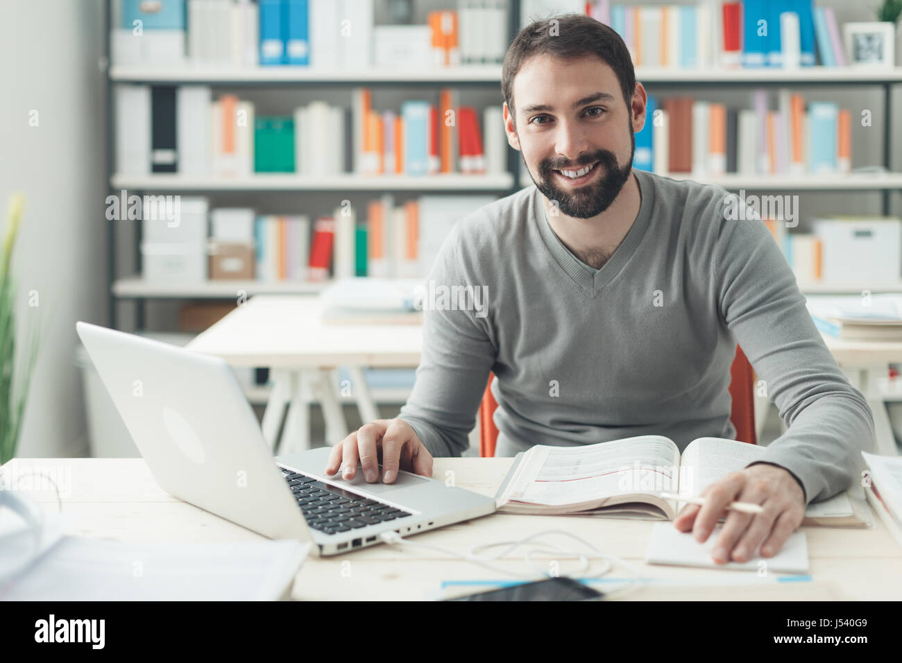 Junger hübscher Mann am Schreibtisch sitzen, ein Buch und Verbindung zum Internet mit einem laptop Stockfoto