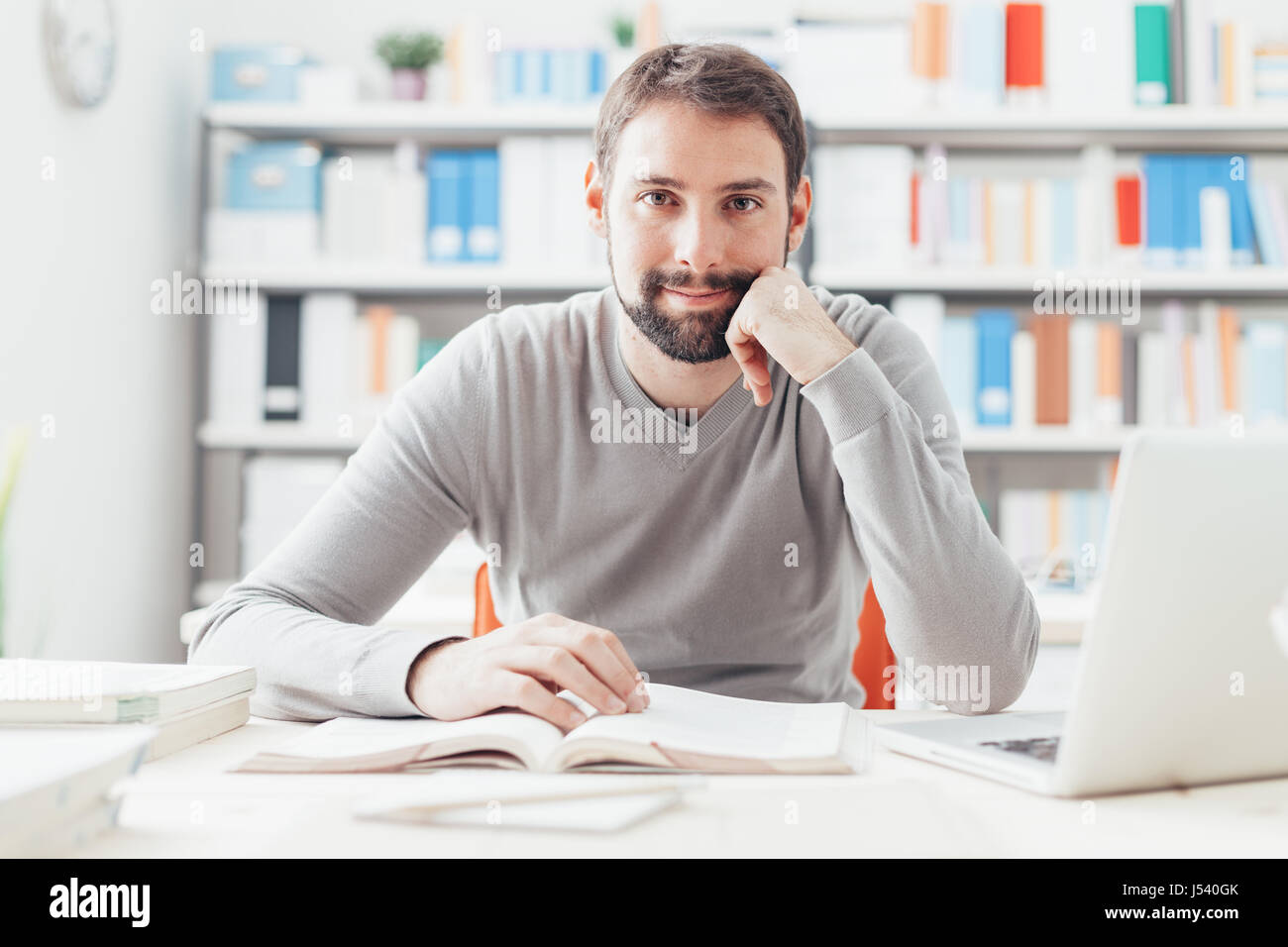 Lächelnde junge Mann, der im Büro arbeitet, ist er ein Buch zu lesen und einen Laptop verwenden Stockfoto