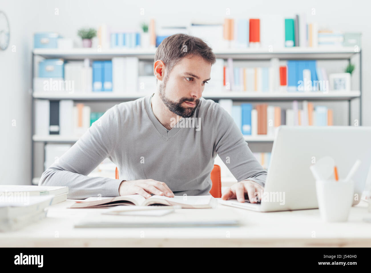 Junger hübscher Mann am Schreibtisch sitzen, ein Buch und Verbindung zum Internet mit einem laptop Stockfoto