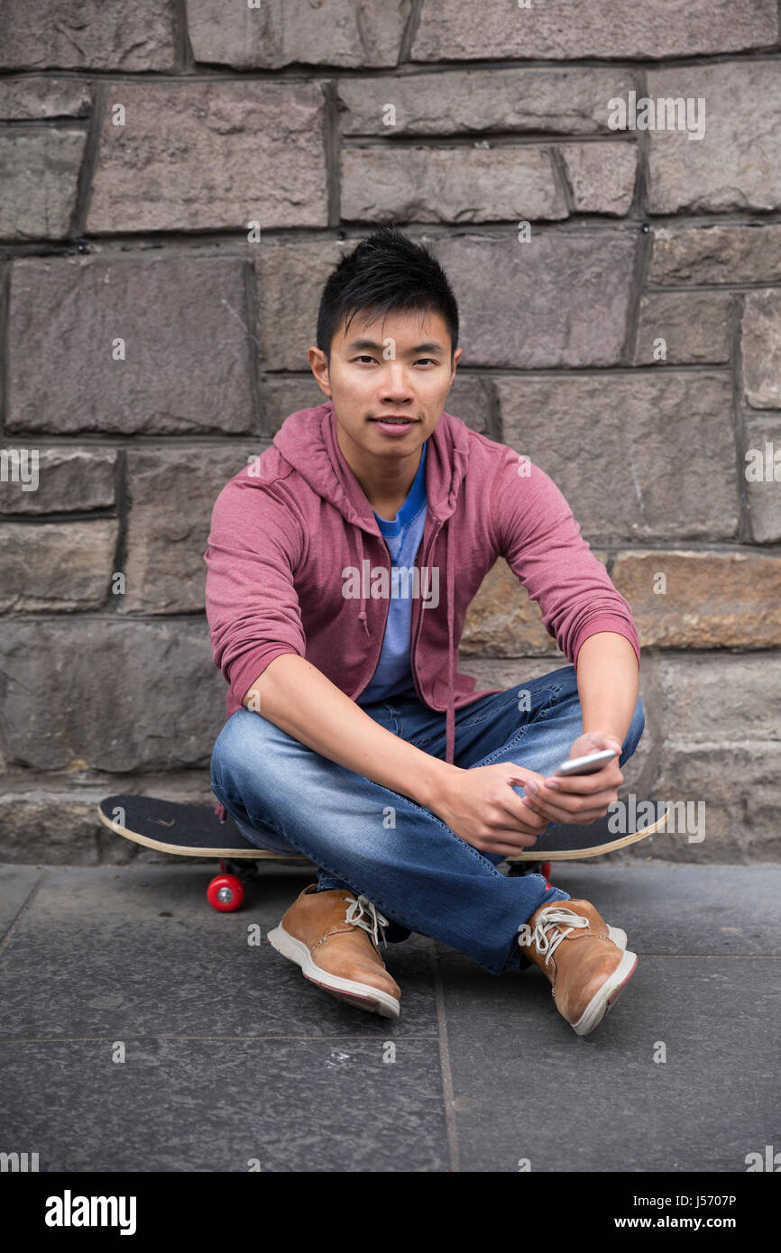 Porträt von einem asiatischen Mann mit seinem Smartphone und hält seinem Skateboard. Im Stadtstraße stehen im Freien. Stockfoto