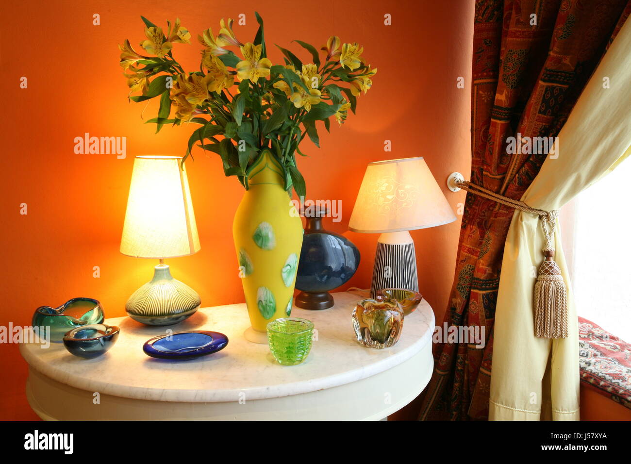 Verschiedene Arten von Glas Ornamente und Lampen mit einer Vase mit Blumen auf einem Marmor tisch Neben einem Vorhang Stockfoto