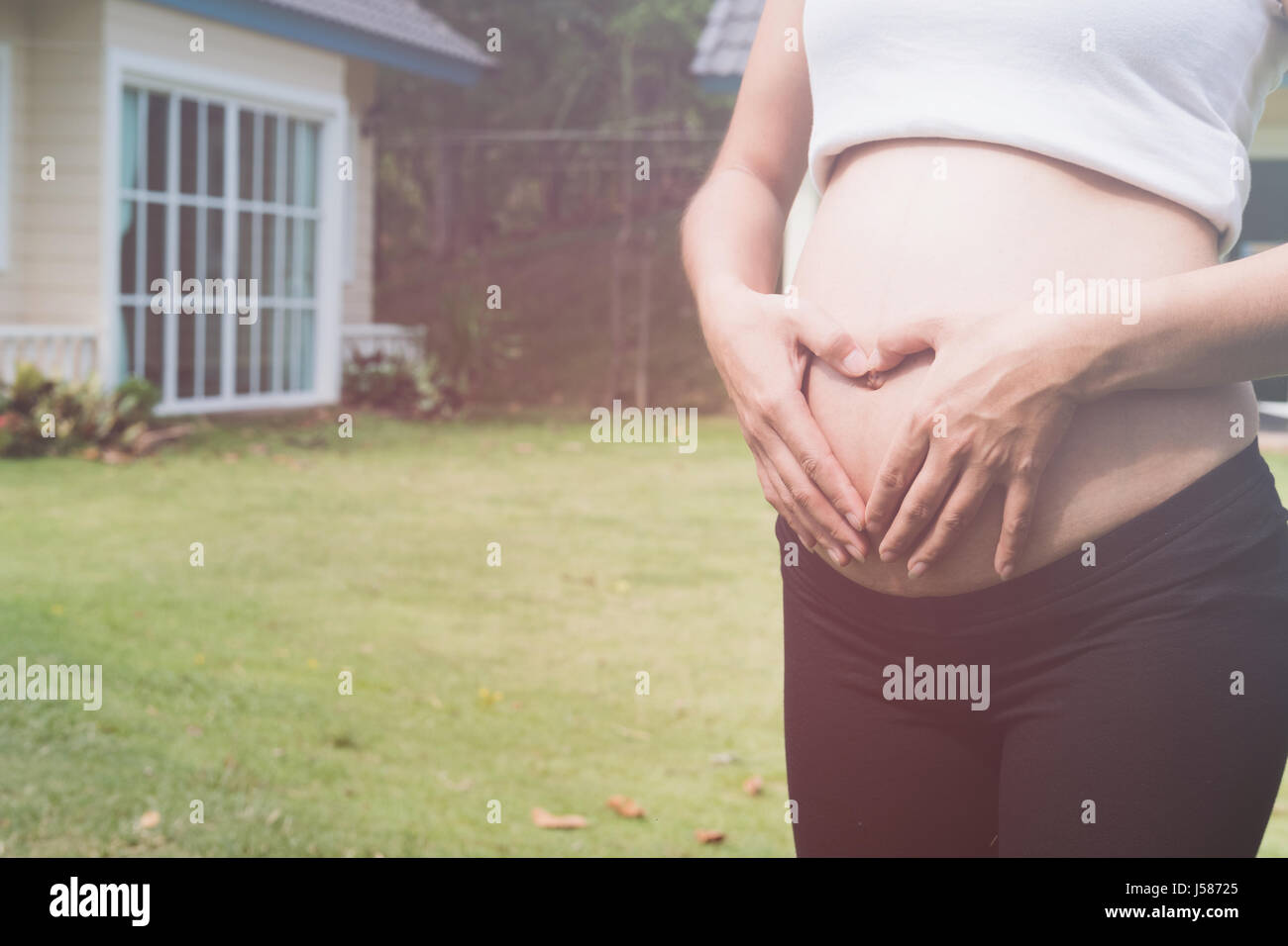 junge asiatische schwangere Frau die Form Herzschild auf ihrem Bauch vor ihrem Haus.  Schwangerschaft, Mutterschaft Bauch Pflegekonzept Stockfoto