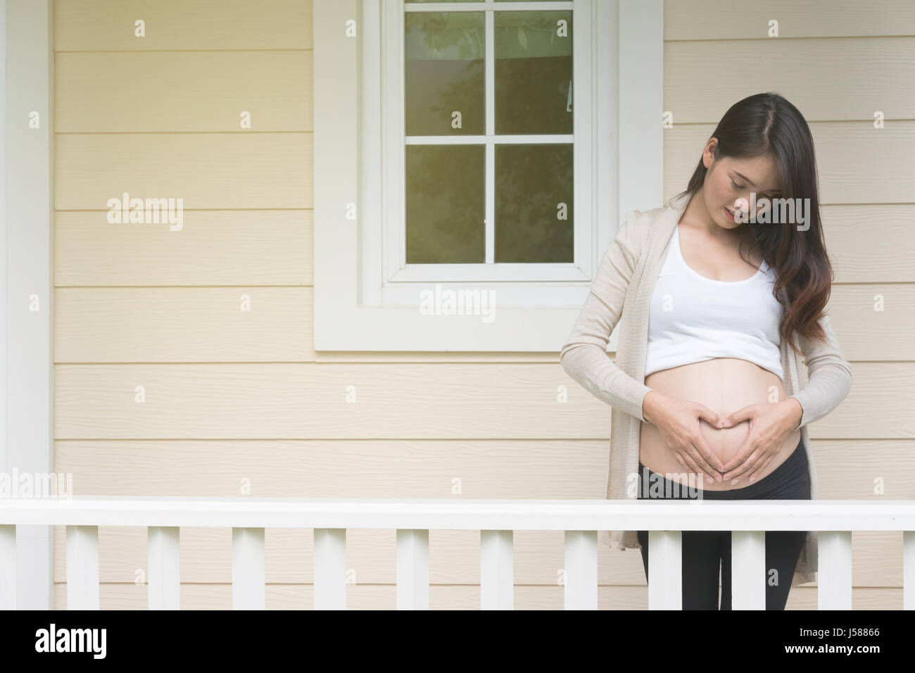 junge asiatische schwangere Frau die Form Herzschild auf ihrem Bauch vor ihrem Haus.  Schwangerschaft, Mutterschaft Bauch Pflegekonzept Stockfoto