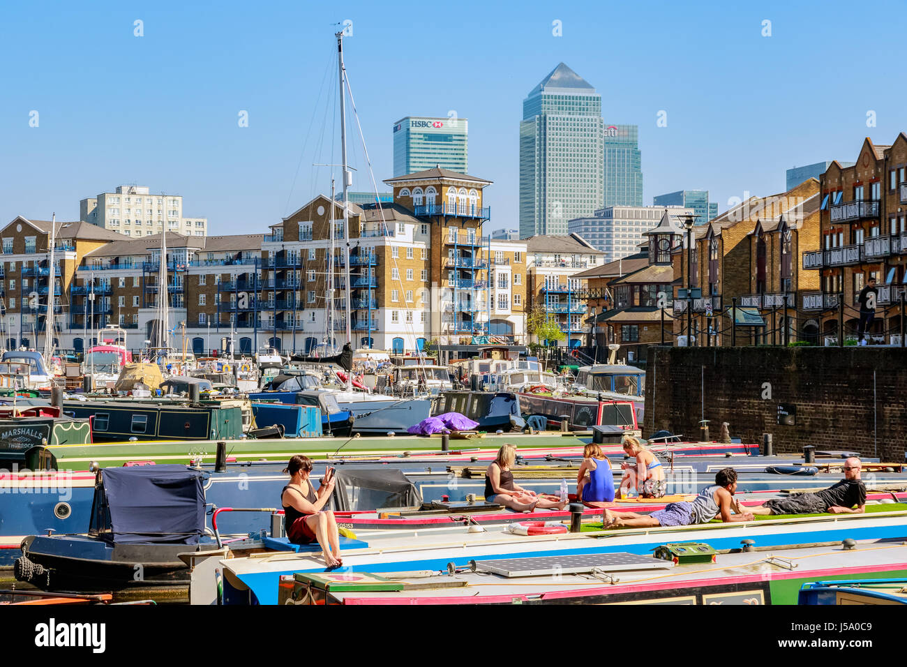 London, UK - 8. April 2017 - Leute zum Sonnenbaden auf den Booten vertäut am Limehouse Bassin mit Canary Wharf im Hintergrund Stockfoto