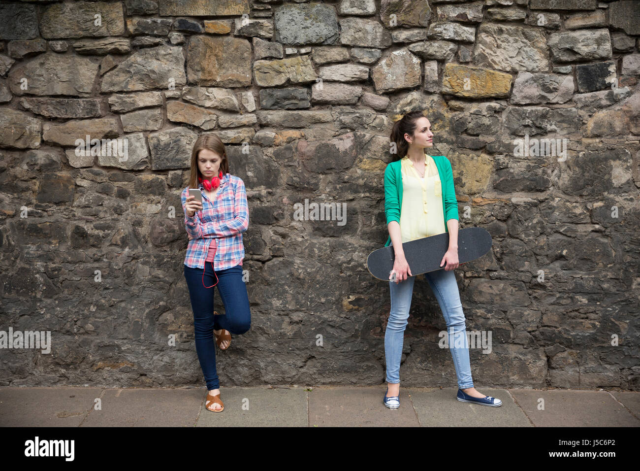 Zwei modische Frauen eine Wand gelehnt, mit einem Skateboard. auf der Suche von Kamera weg. Stockfoto