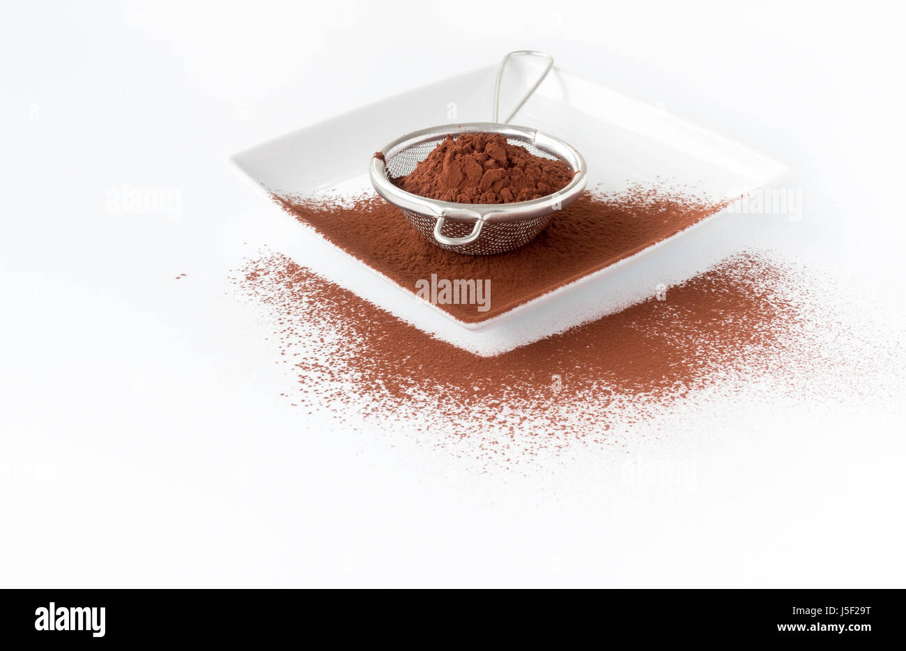 Nahaufnahme von einem Metallsieb mit Kakaopulver auf einem weißen Quadrat Porzellanteller gefüllt. Weißen Hintergrund mit Textfreiraum, flachen Schärfentiefe und sel Stockfoto