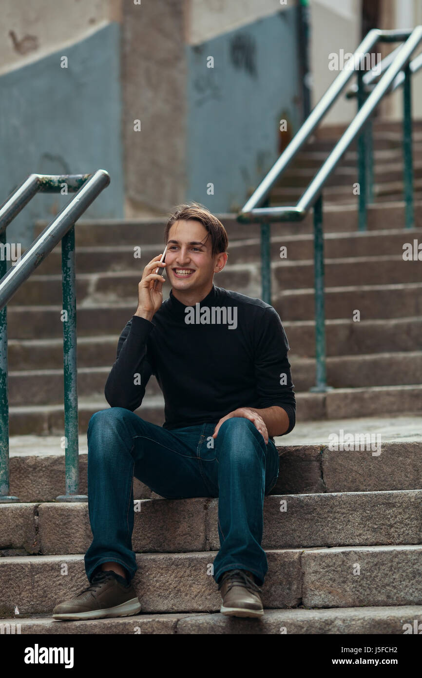 Hübscher junger Mann telefonieren mit Handy beim Sitzen auf Steinstufen im Freien. Stockfoto
