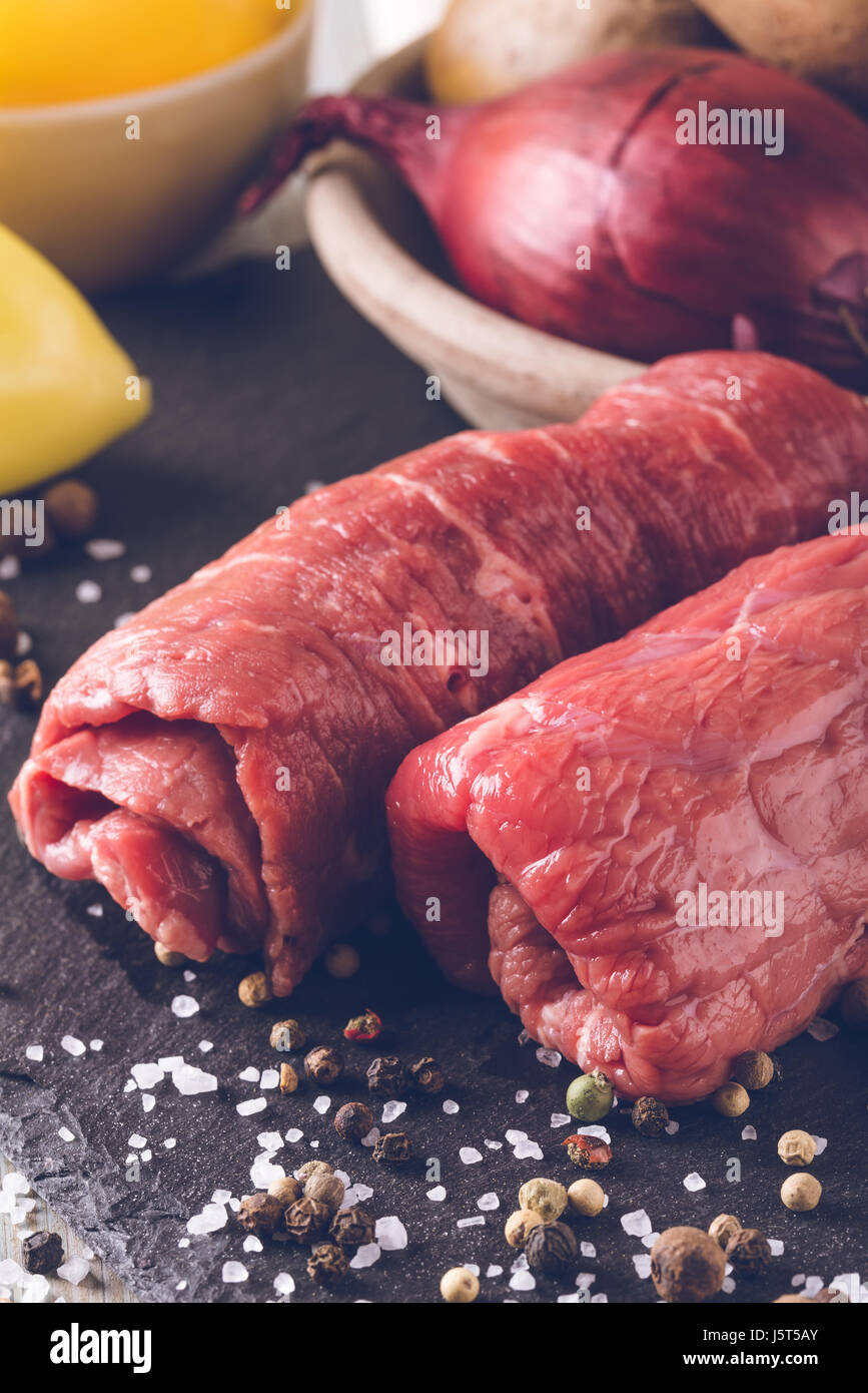 Vertikale Vintage Foto mit Rindfleisch rollt. Zwei dünne Scheiben rot rohes Fleisch für Rouladen vorbereitet. Essen auf schwarzem Schiefer mit gelber Paprika Stockfoto