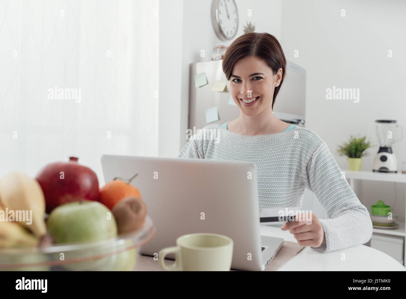 Lächelnde junge Frau mit einem Laptop in der Küche zu Hause verbinden, tut sie, Online-shopping und Kauf von Produkten unter Verwendung einer Kreditkarte Stockfoto