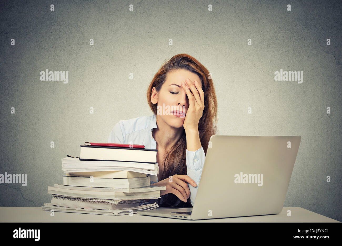 Zuviel Arbeit müde verschlafene junge Frau sitzt an ihrem Schreibtisch mit Büchern vor Laptopcomputer isoliert graue Wand Office-Hintergrund. Terminkalender Stockfoto