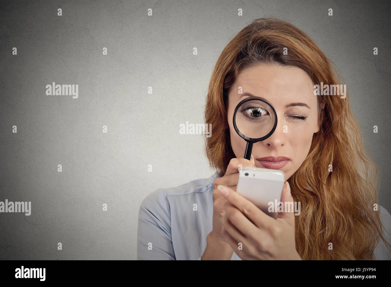 Neugierig. Blick durch die Lupe auf Smartphone-Bildschirm Geschäftsfrau isoliert grau hinterlegt. Menschliches Gesichtsausdruck. Ermittler suchen Stockfoto