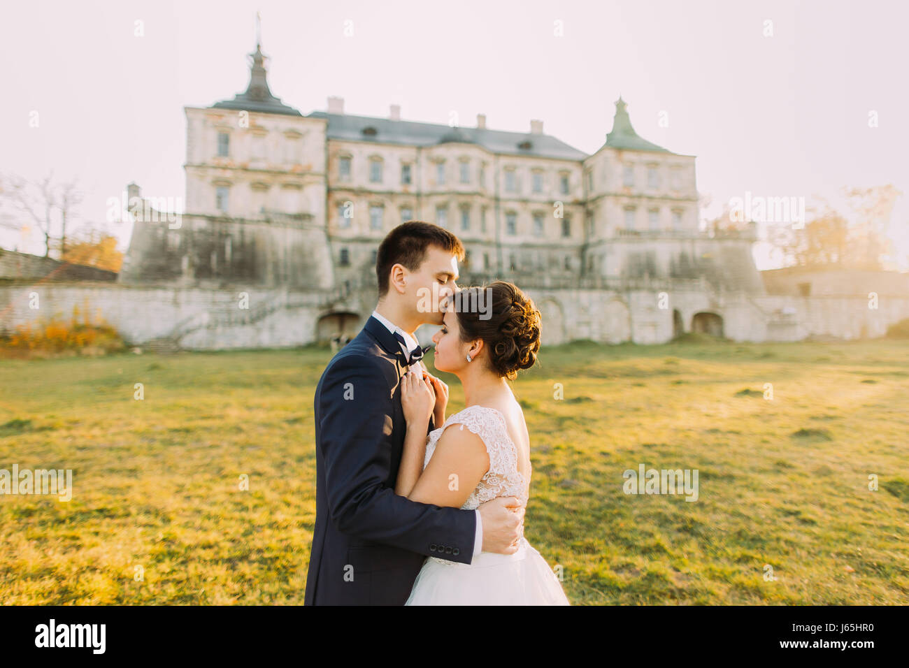 Das horizontale Seite Porträt das glückliche Brautpaar auf dem Hintergrund des alten Palastes. Der Bräutigam ist die Braut während des Sonnenuntergangs in der Stirn küssen. Stockfoto