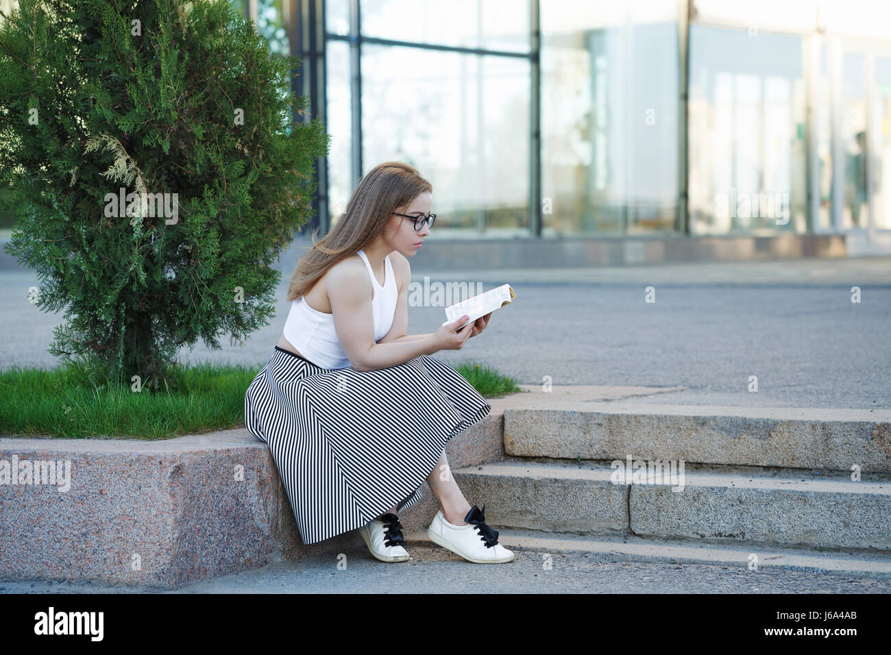 Junge attraktive Hipster Mädchen liest ein Buch auf den Stufen. Sie ist in Gläser, Top, Rock und Turnschuhen bekleidet. Studentin ist selbst erziehen Stockfoto