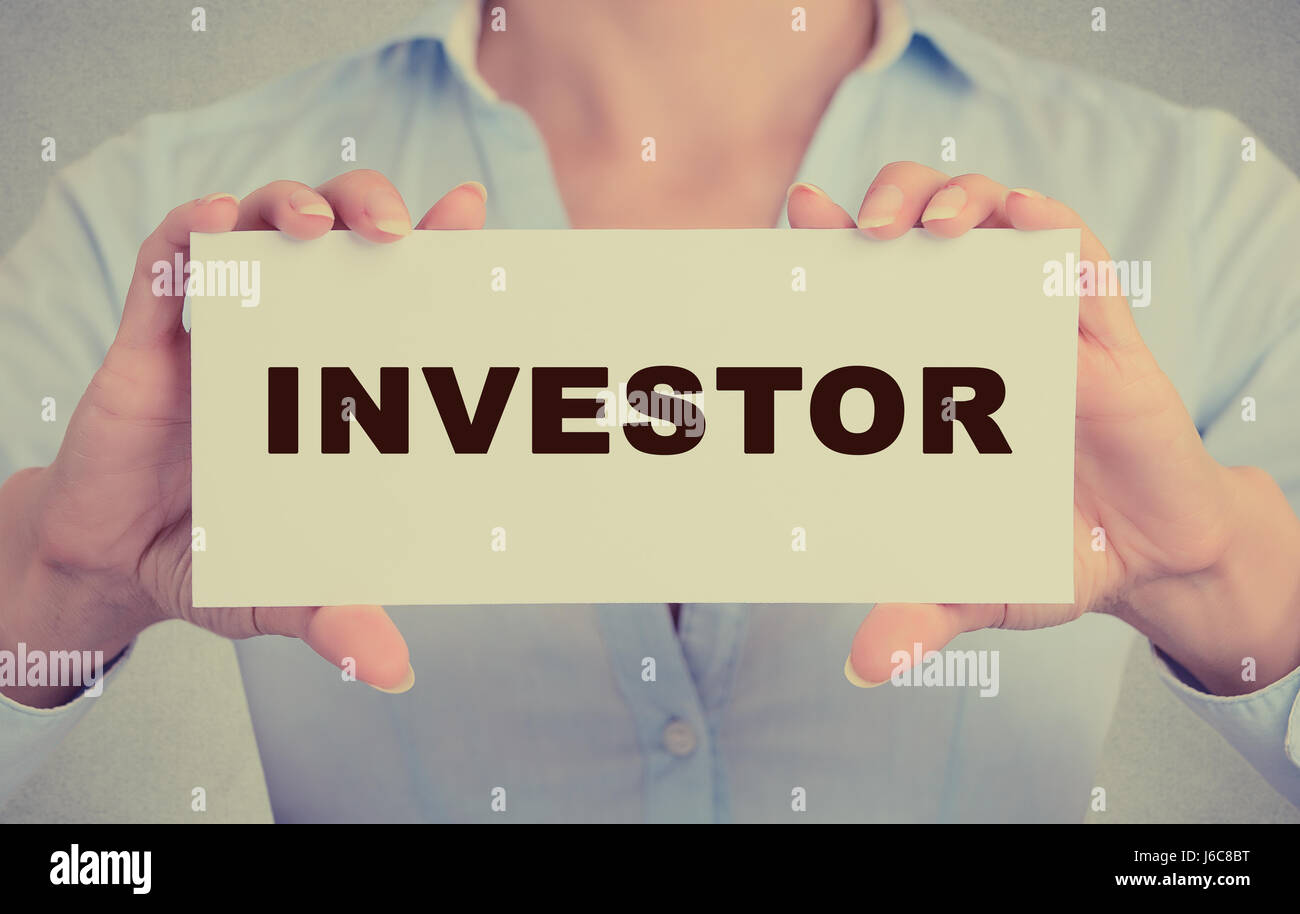 Geschäftsfrau Hände halten weiße Karte Zeichen mit Investor SMS auf graue Wand Büro Hintergrund isoliert. Retro-Instagram Stil Bild. Finanzielle Stockfoto