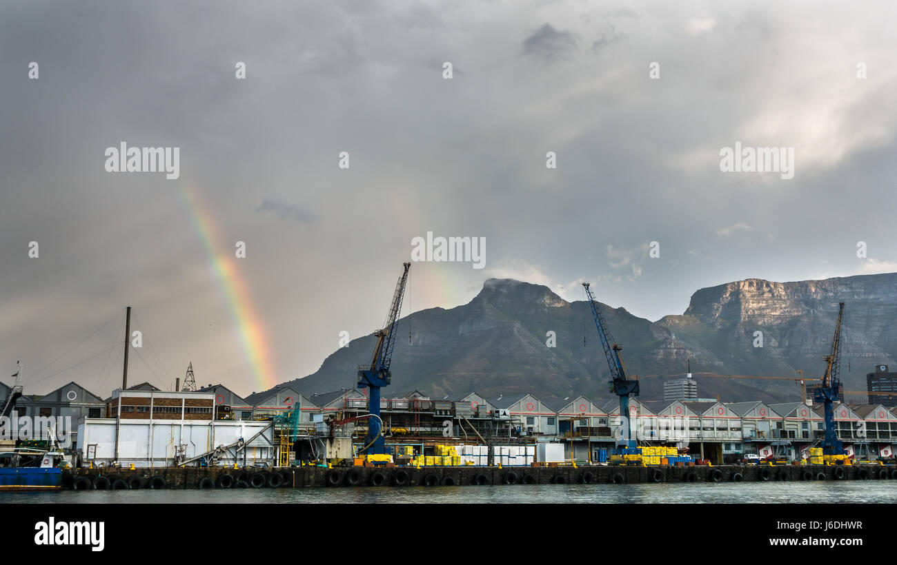 Tafelberg und Devil's Peak mit doppeltem Regenbogen hinter Kranichen im Hafen und dunklem stürmischen Himmel, Victoria Basin, Hafen von Kapstadt, Südafrika Stockfoto