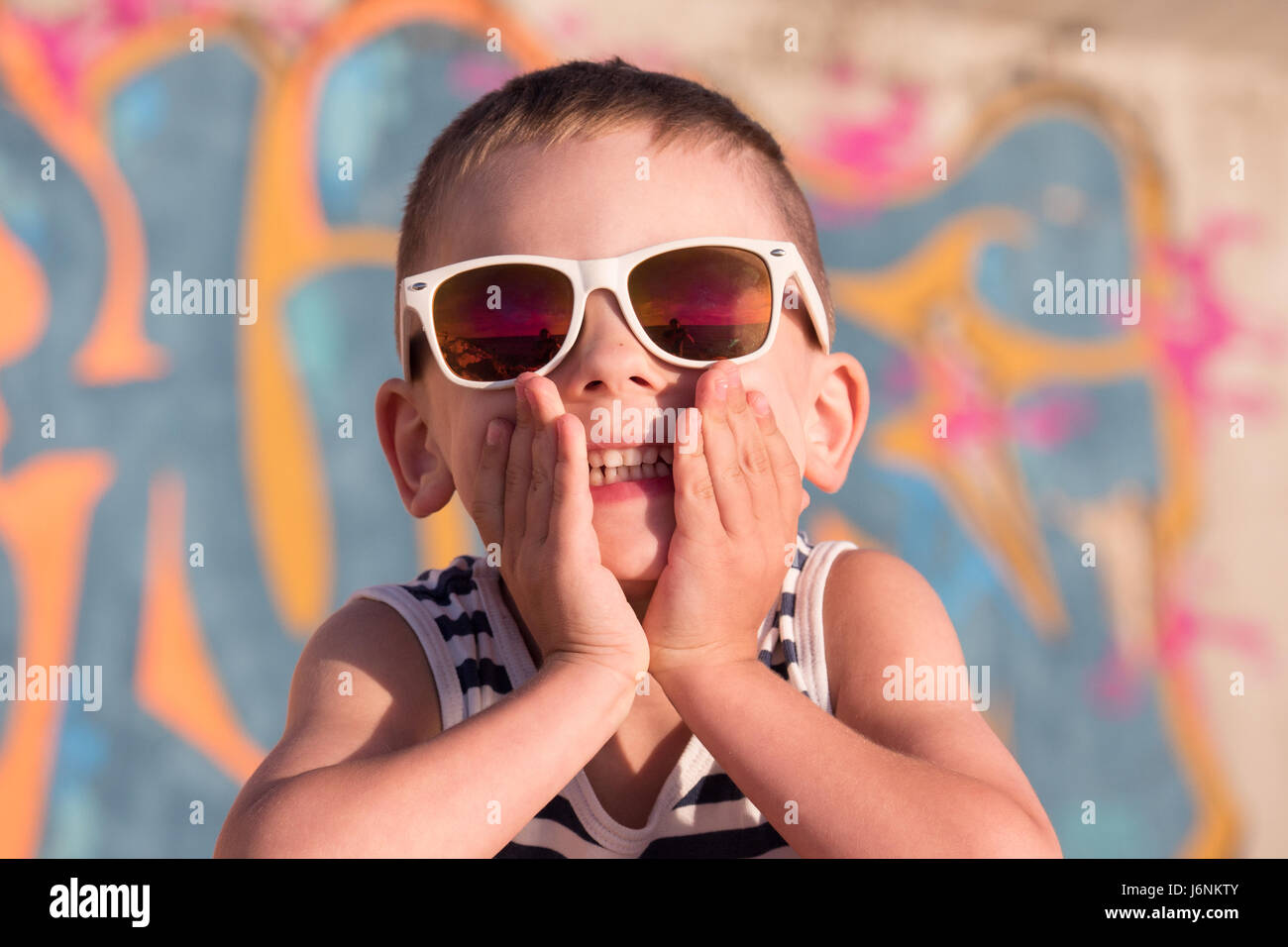 lächelnde Knabe tragen Sonnenbrillen und gestreiften Hemd geschlossen seinen Mund mit den Händen auf Graffiti Hintergrund Stockfoto