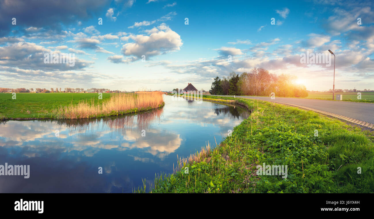 Gebäude und Bäume in der Nähe der Wasserkanal bei Sonnenaufgang in Niederlande. Bunte blauer Himmel mit Wolken. Sommerlandschaft. Ländliches Motiv. Bewölkter Himmel reflektiert Stockfoto