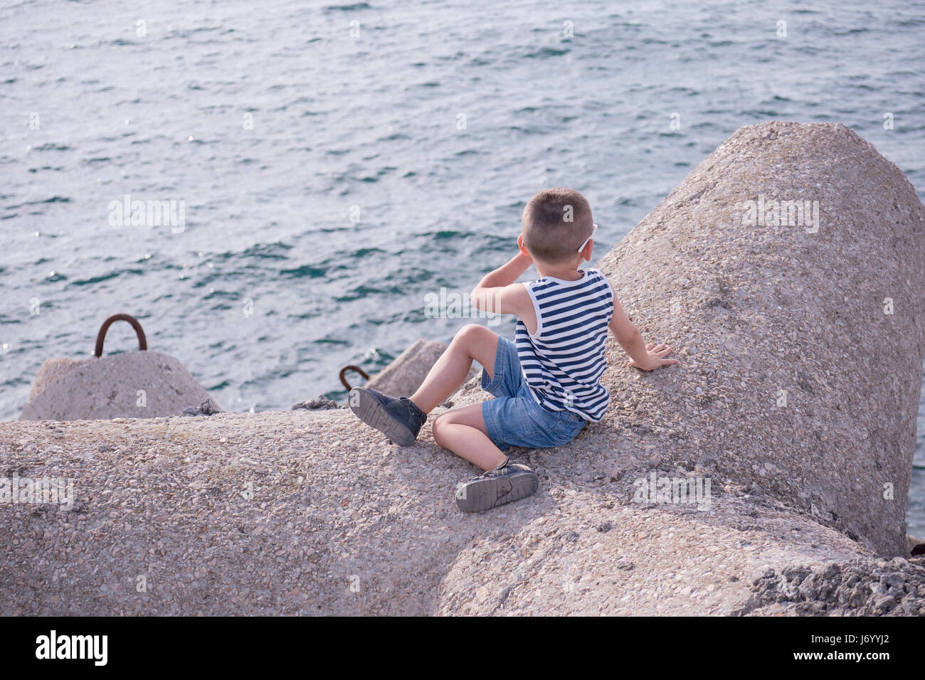 gesunde junge gekleidet in ein gestreiftes Shirt und Jeans-Shorts sitzen auf konkrete Wellenbrecher am Meer Stockfoto