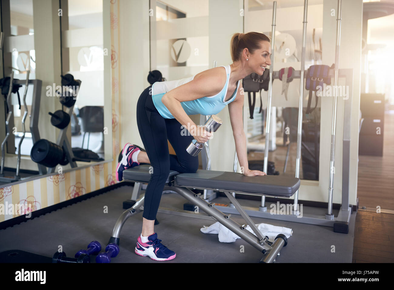 Frau mittleren Alters mit einer Hantel Gewicht in einem modernen Fitnessstudio mit Spiegel und eine Vielzahl von Geräten, die über eine Bank als She Exerc nach vorne lehnen Stockfoto