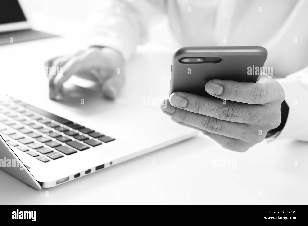 Nahaufnahme von Geschäftsmann mit Handy und Laptop-Computer auf weißen Schreibtisch im modernen Büro, schwarz / weiß Stockfoto