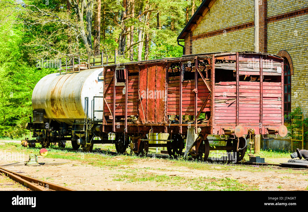 Rote Vintage Holz Güterwagen stehen verlassen auf Gleis außerhalb der gelbe Backsteinbau mit chemischen Auto und Wald im Hintergrund. Stockfoto