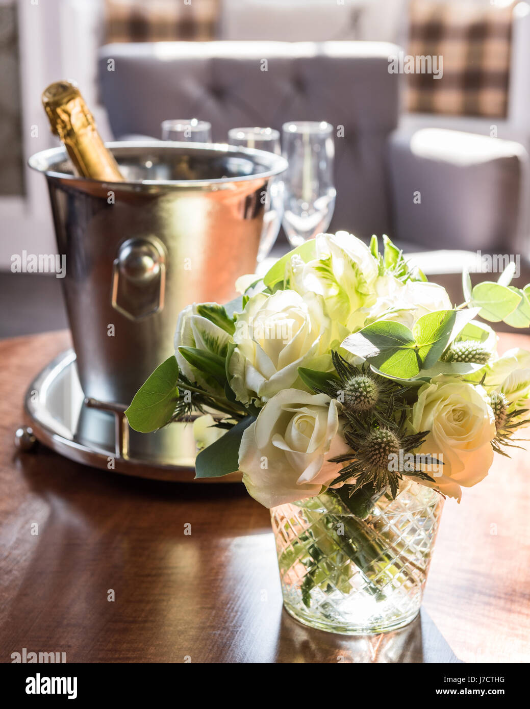 Eine posey von weißen Rosen und Disteln in Glasvase auf Tisch mit Champagner und Silber Eimer im Hintergrund Stockfoto