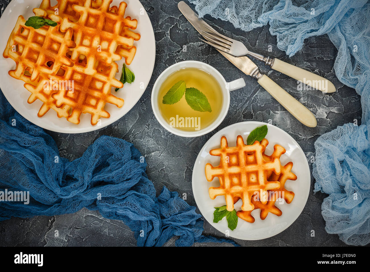 Zwei Platten mit belgischen Waffeln garniert mit Minze, Glas Tasse grüner Tee mit Minze, Gabel mit Messer auf grauem Hintergrund. Ansicht von oben Stockfoto