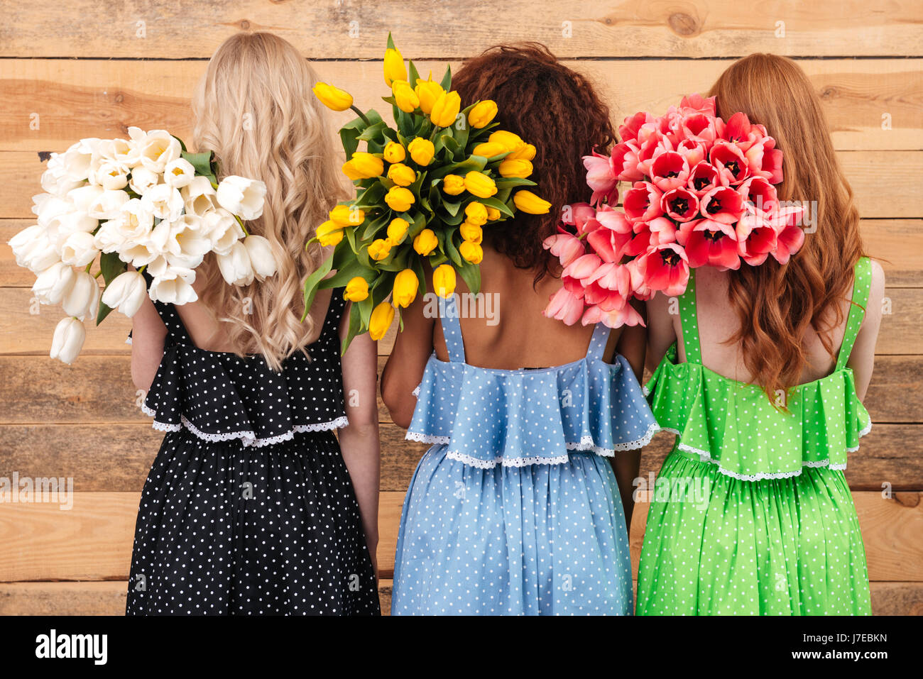 Rückansicht der drei Frauen in Kleidern Stand in der Nähe der Wand und hält Blumenstrauss Stockfoto