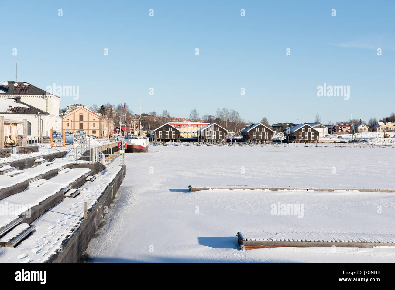 Hudiksvall gefrorener Hafen mit Booten und traditionellen Holzlagern unter klarem blauem Winterhimmel in einer skandinavischen Küstenstadt Stockfoto