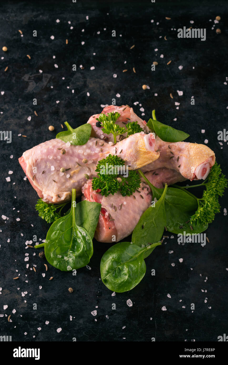 Vertikale Foto von rohem Hühnerfleisch Beine. Drei Stücke von Fleisch sind auf dunkle Vintage Tablett gelegt. Grüne Kräuter wie Petersilie und Spinat sind um mit verschüttet. Stockfoto