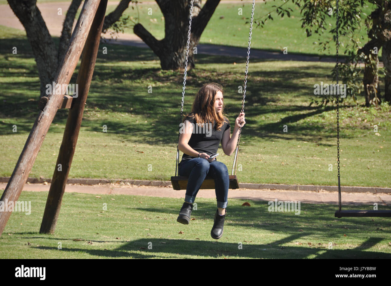 hübsche blonde Teen in Jeans und kurzen Ärmeln und Stiefeln setzt auf Holz und Kette Schaukel in einem Park mit Schatten und Bäume im Hintergrund. Stockfoto