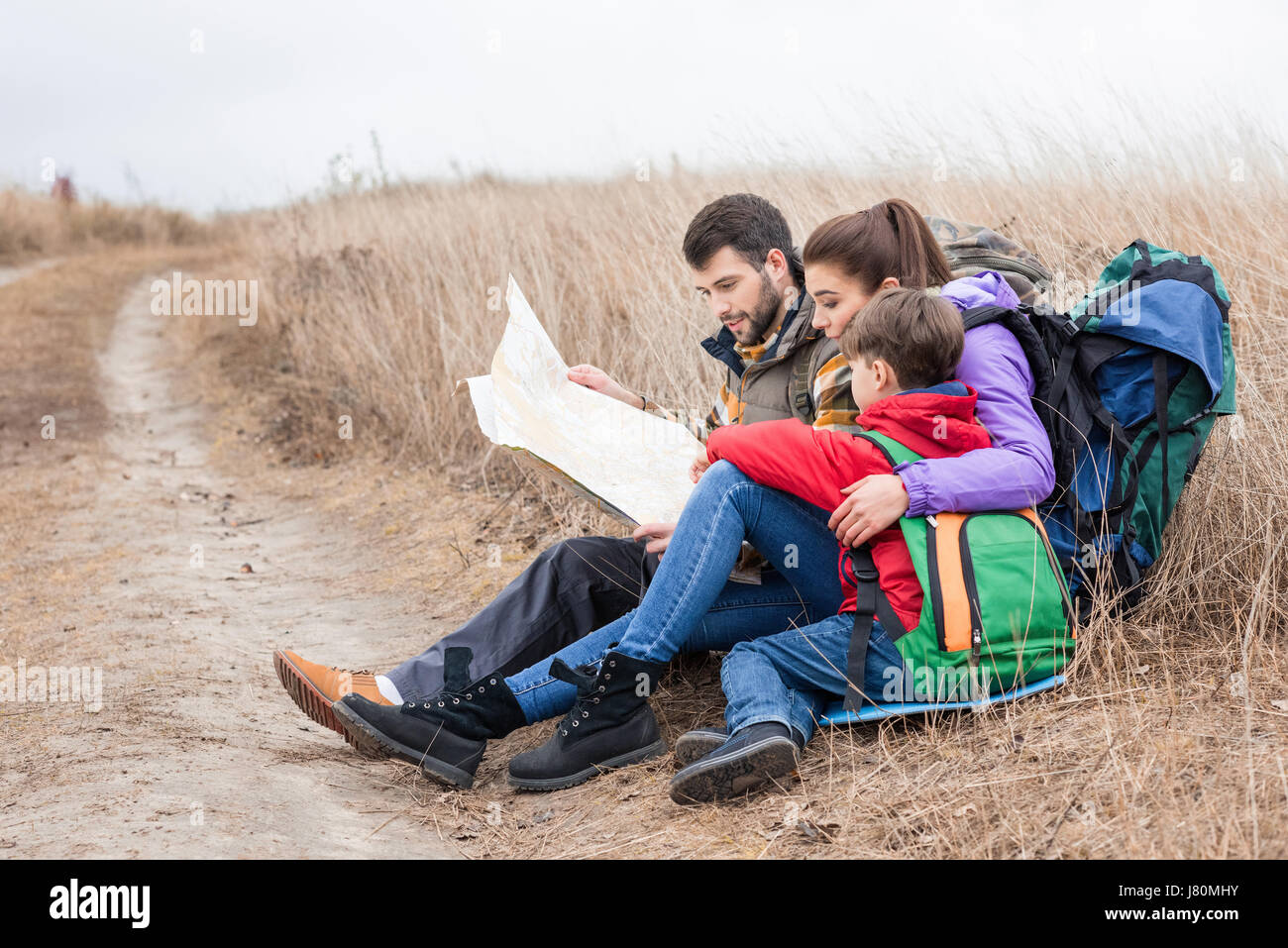 Junge Familie mit Rucksäcken auf dem Rasen sitzen und Blick auf Karte in Landschaft am Herbsttag Stockfoto