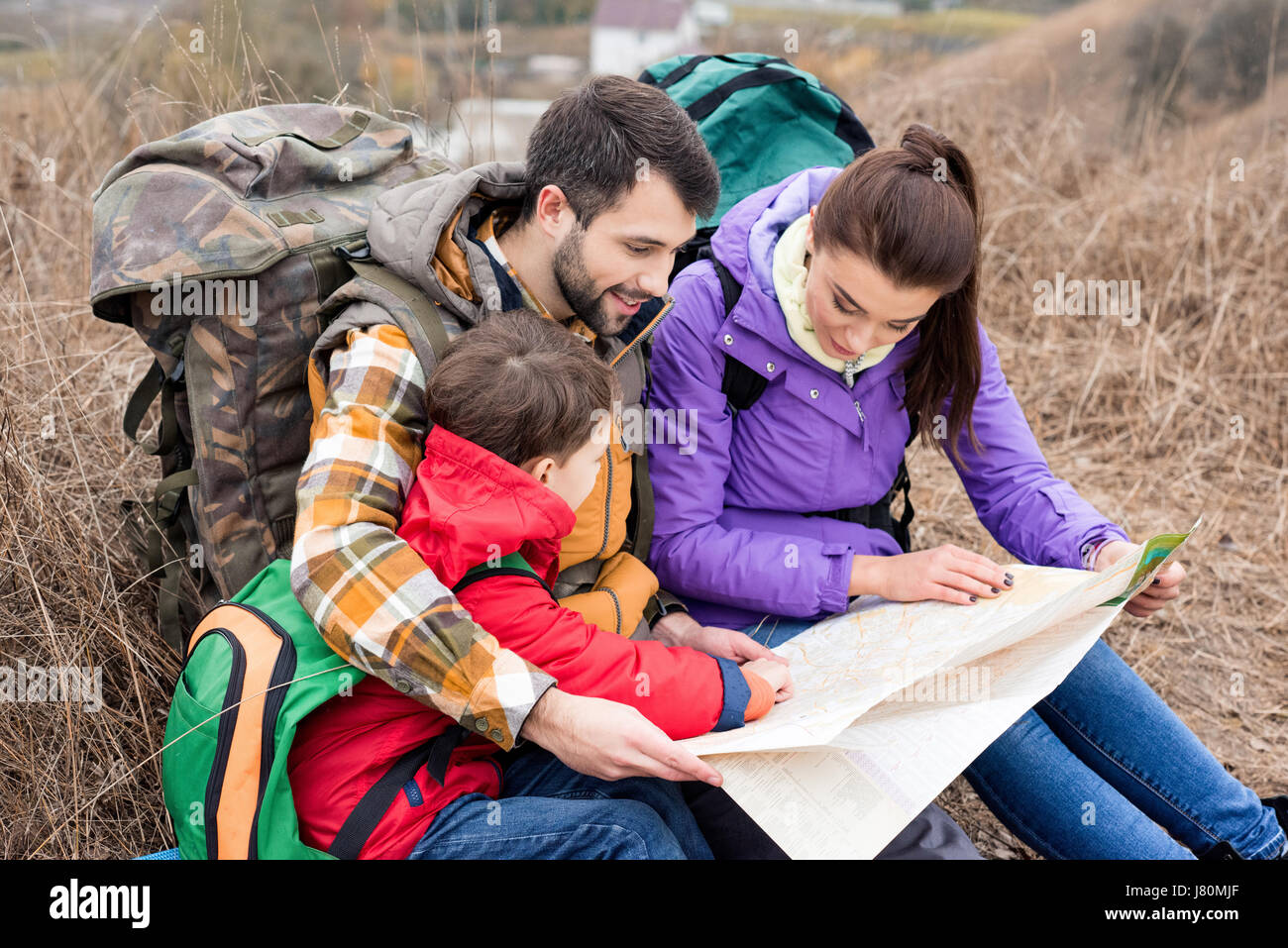 Junge Familie mit Rucksäcken auf dem Rasen sitzen und Blick auf Karte in Landschaft am Herbsttag Stockfoto