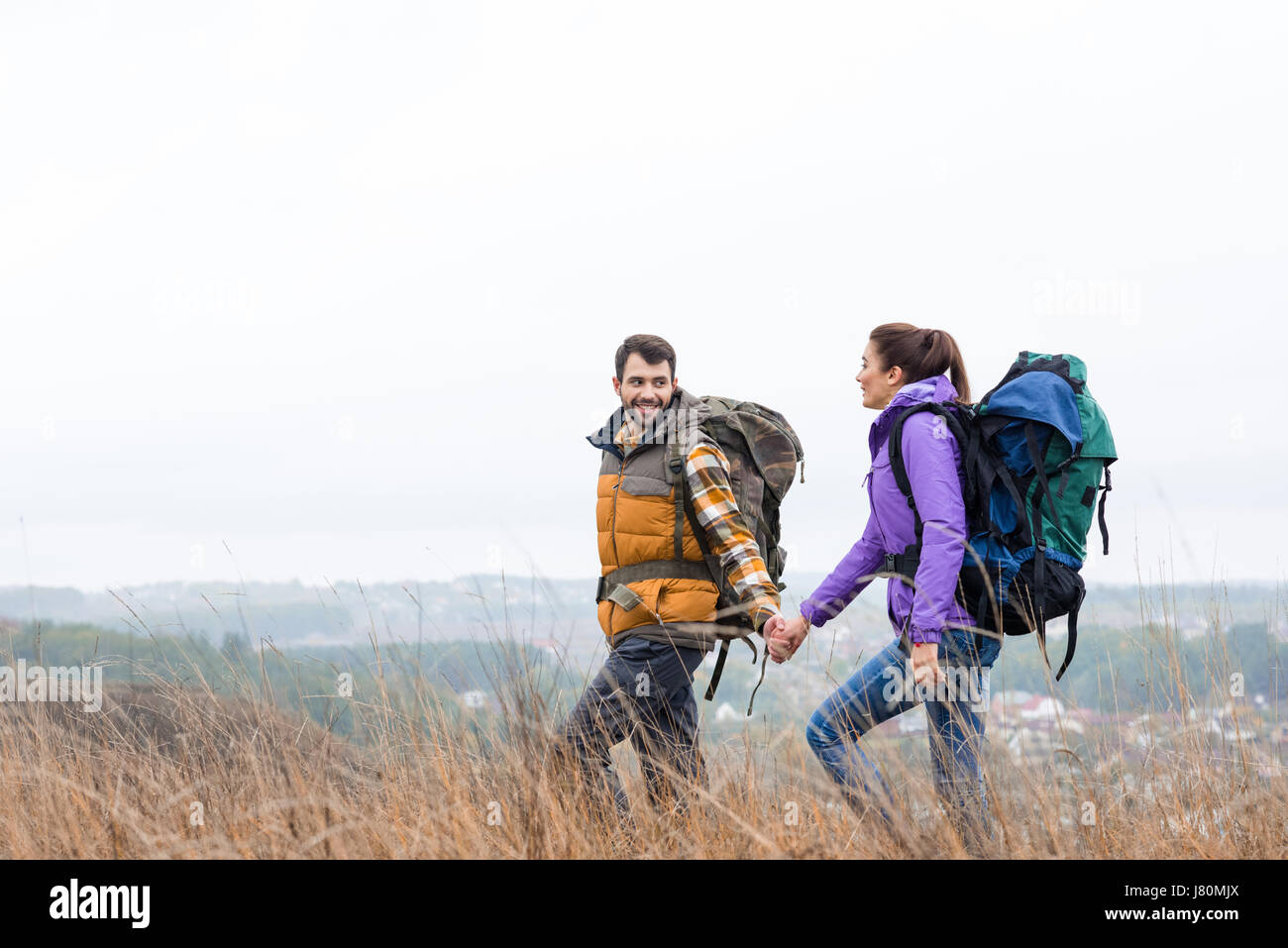 Junge, lächelnde paar mit Rucksäcken, Hand in Hand und Fuß hohen Gras im ländlichen Raum Stockfoto