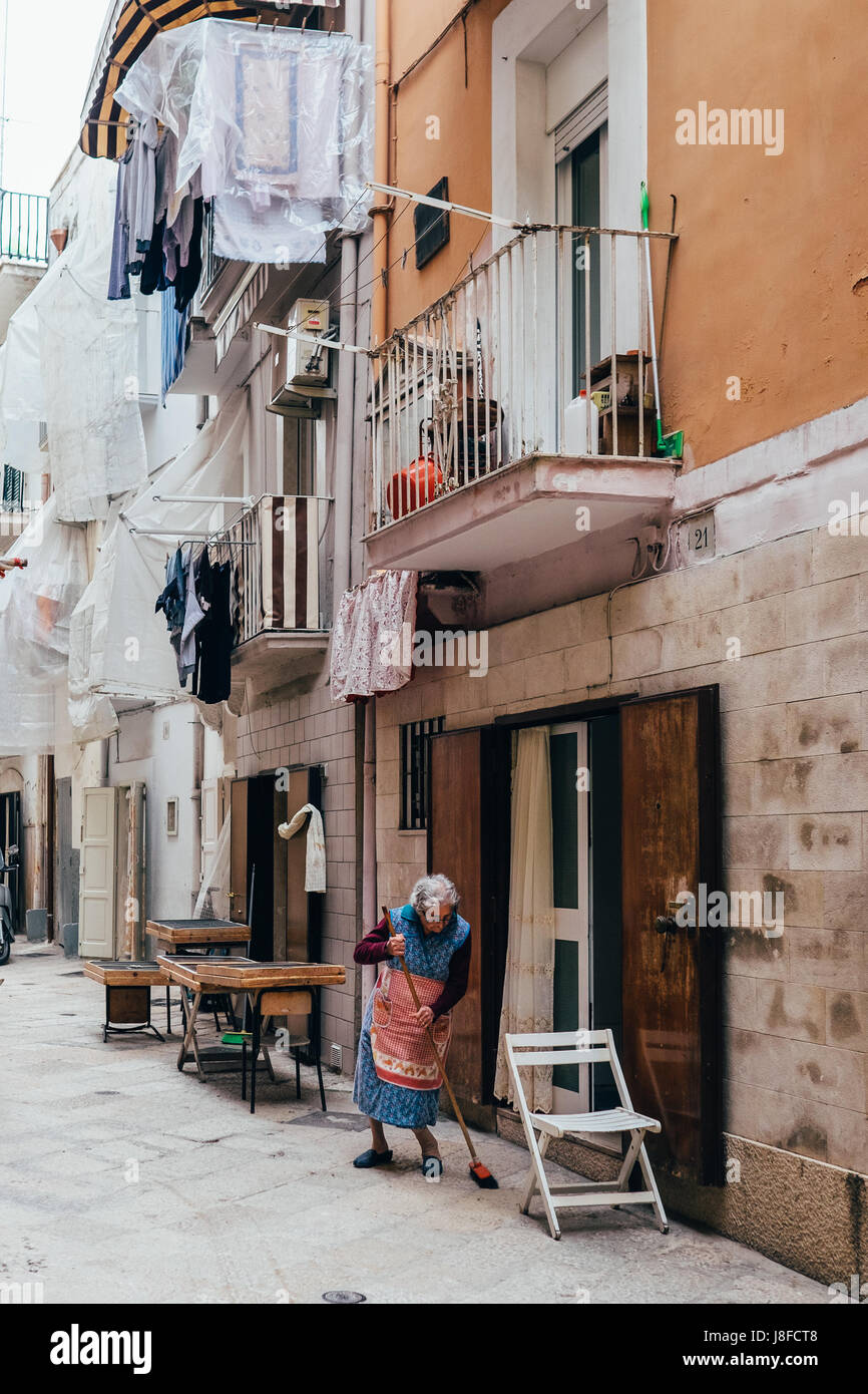 Eine ältere Frau fegt die Straße, während Schalen mit traditionellen Orecchiette Nudeln trocknen in der Nähe in Bari Vecchia, Apulien, Italien Stockfoto