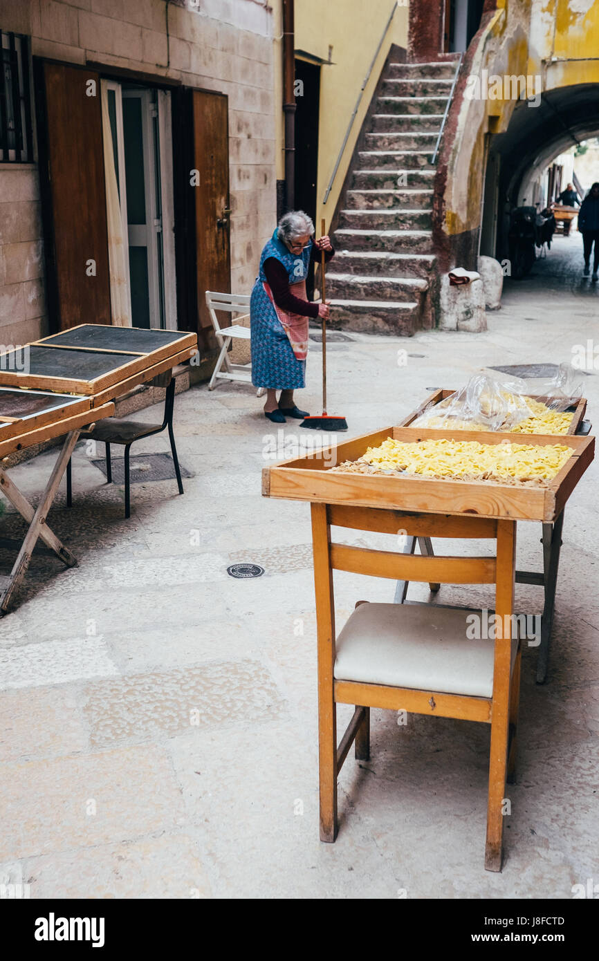 Eine ältere Frau fegt die Straße, während Schalen mit traditionellen Orecchiette Nudeln trocknen in der Nähe in Bari Vecchia, Apulien, Italien Stockfoto