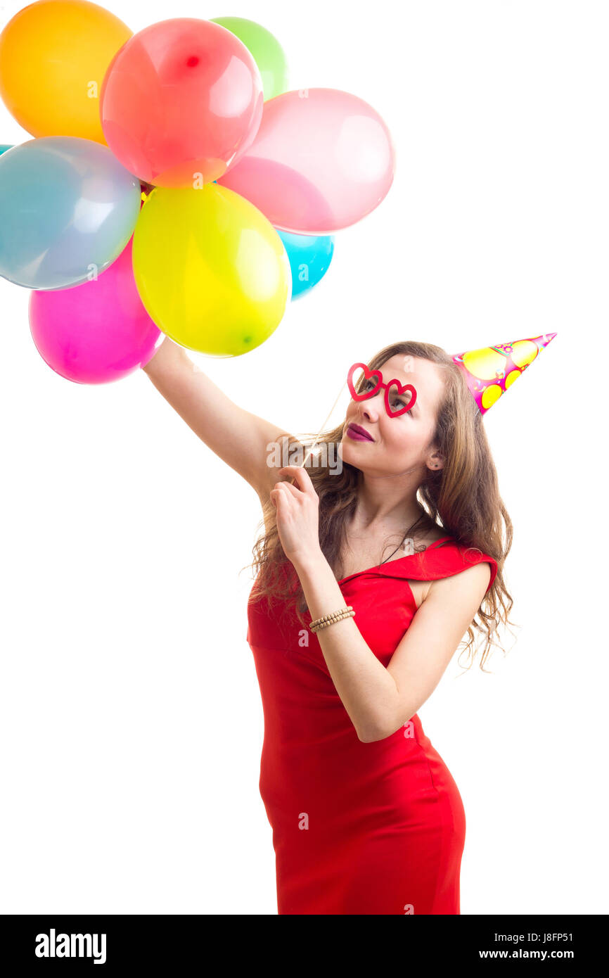 Junge, elegante Frau mit lange kastanienbraunes Haar im roten Kleid mit Hut hält viele bunten Luftballons und Karte Stick Brille auf weißen Hinterg feiern Stockfoto