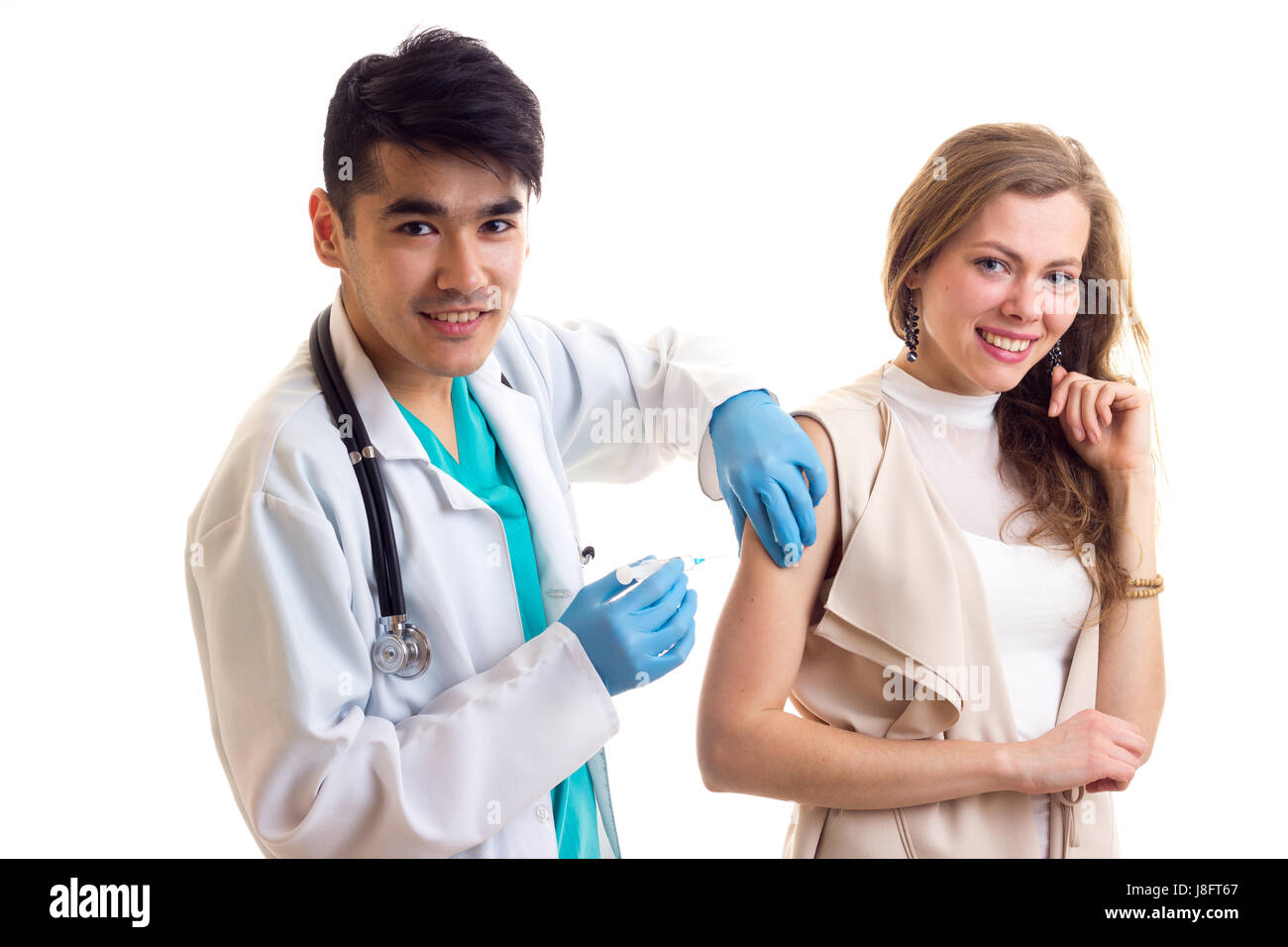 Junge Smartlooking Arzt mit dunklem Haar im weißen Kleid mit Stethoskop und blaue Handschuhe machen eine Injektion von junge lächelnde Frau mit langen braunen hai Stockfoto