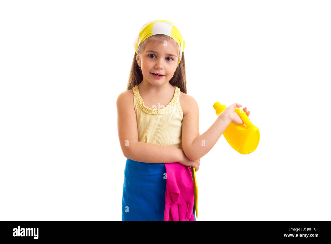 Lustige kleine Mädchen mit langen kastanienbraunes Haar in gelbes Hemd und blaue Schürze mit rosa Handschuhe und gelbes Halstuch gelb Duster und gelbe Flasche halten Stockfoto