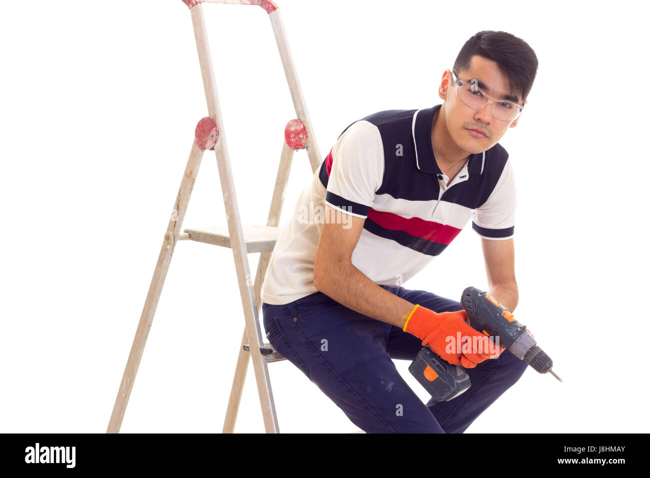 Junger attraktiver Mann mit schwarzen Haaren in weißen und blauen T-shirt mit orange Handschuhe und Schutzbrille grau Akkuschrauber halten und sitzen Stockfoto