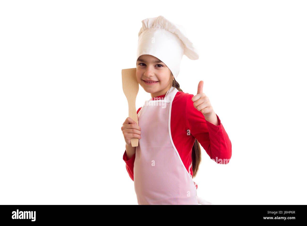 Hübsche Mädchen im roten Hemd mit weißer Schürze und Hut hält Küche Spachtel und Daumen zeigt auf weißem Hintergrund im studio Stockfoto