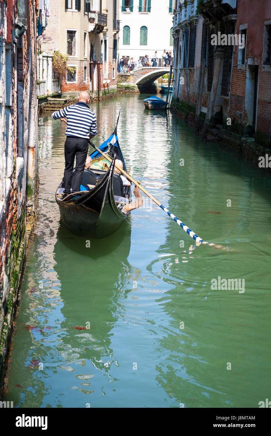 Reizvolle Aussicht auf einen kleinen Kanal in Venedig, Italien, mit Gondoliere im traditionellen gestreiften Hemd Gondel an grünen Gewässern manövrieren Stockfoto