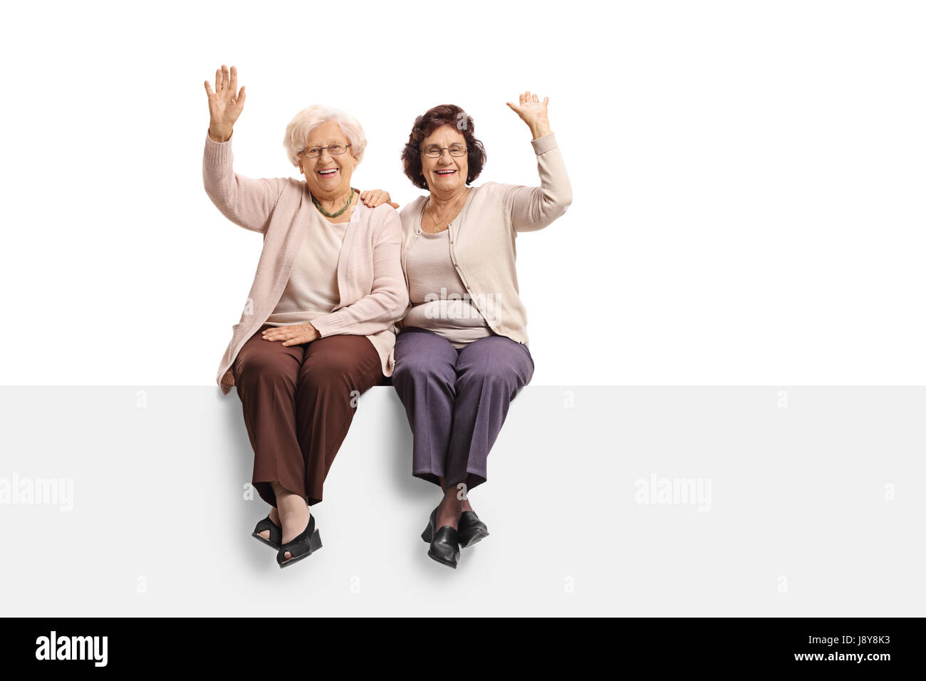 Zwei fröhliche Reife Frauen, die auf einer Tafel sitzen und winken auf weißem Hintergrund isoliert Stockfoto
