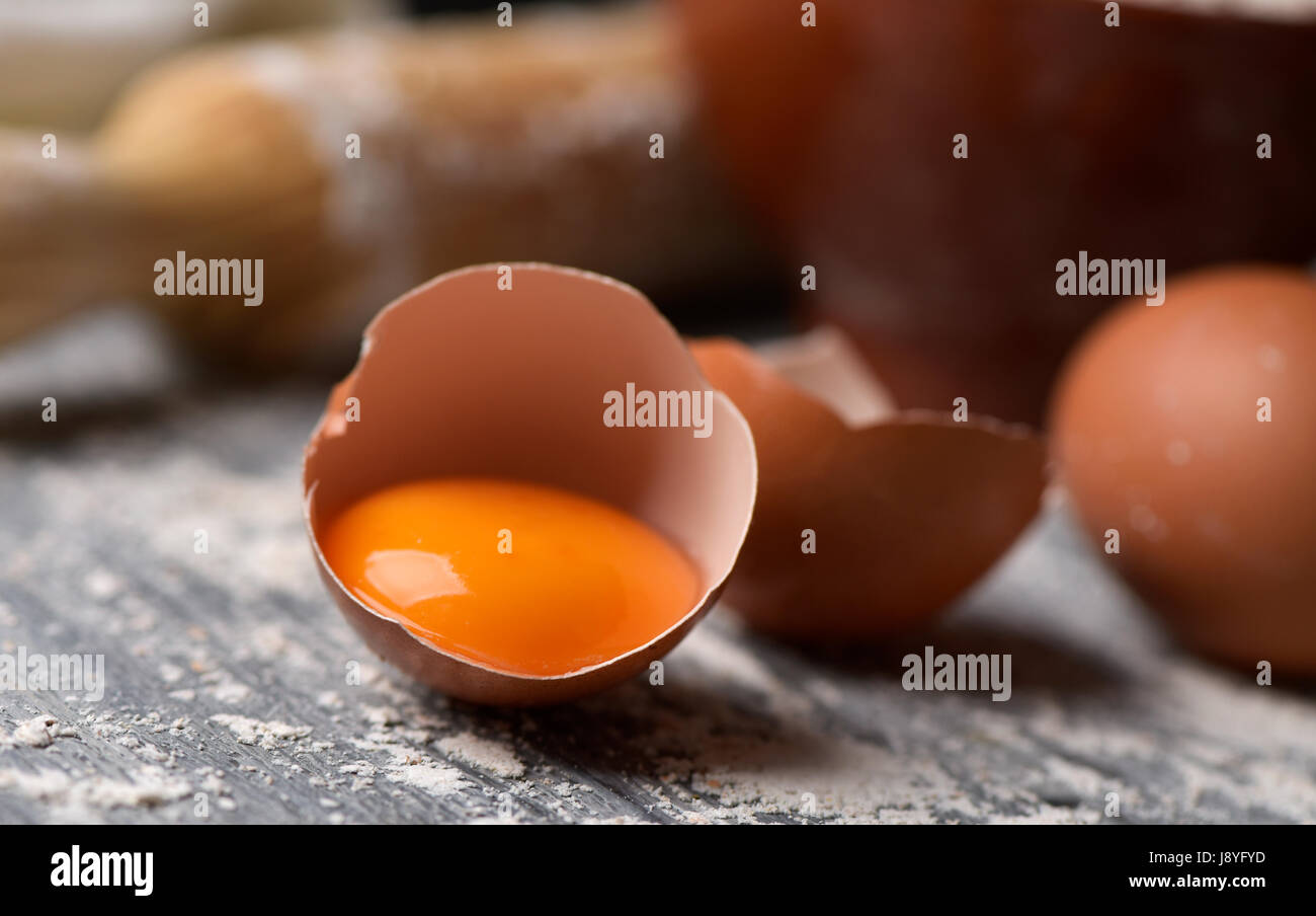 Nahaufnahme von einem gerissenen Ei, ein ganzes Ei und einem hölzernen Nudelholz auf einem rustikalen Tisch mit Mehl bestreut Stockfoto