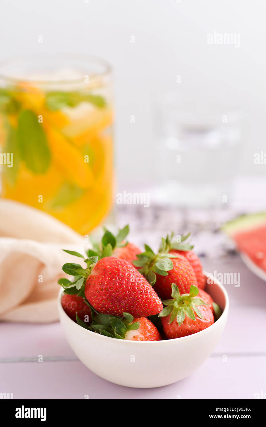 Nahaufnahme von einer weißen Keramikschale mit Erdbeeren auf einem Tisch und einem Glas mit kaltem Tee im Hintergrund Stockfoto